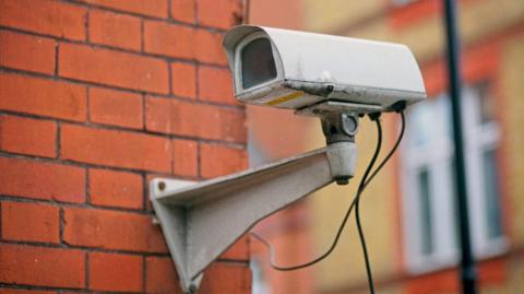 A white, rectangular, CCTV camera mounted on a red brick wall