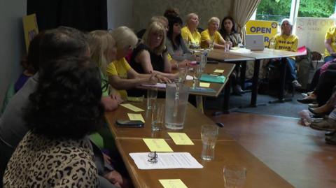 A group of people sat along wooden tables. Some people are wearing yellow t-shirts with Save Our Ward written on in blue