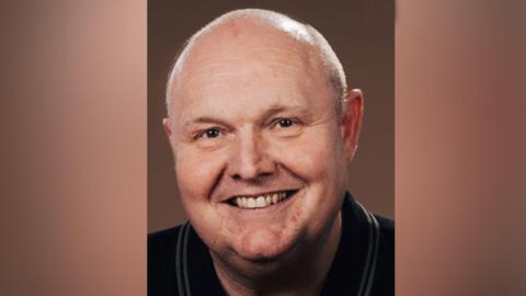 A bald man with brown eyes, smiling towards the camera. 