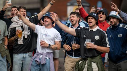 A group of Oasis fans in Cardiff with their arms in the air with some holding pints of beer. They are wearing the band's merchandise, including sport shirts with the logo on, along with bucket hats and are standing in a street. 