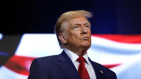 Donald Trump looks ahead with a serious expression on his face. He is wearing a navy suit, white shirt and red tie.