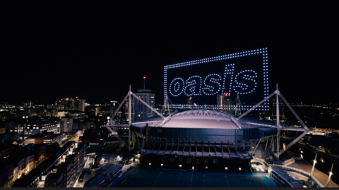 The outside of the Principality Stadium at night with high-rise buildings surrounding it. Above the stadium are purple lights in the sky which spell out Oasis, with two rectangle boxes surrounding the letters.
