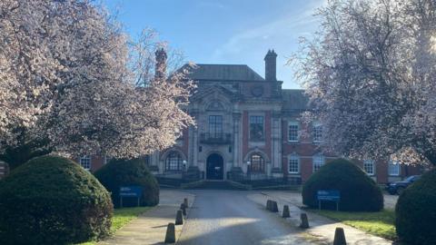 A long driveway leading to a large two storey building in a mixture of stone and red brick