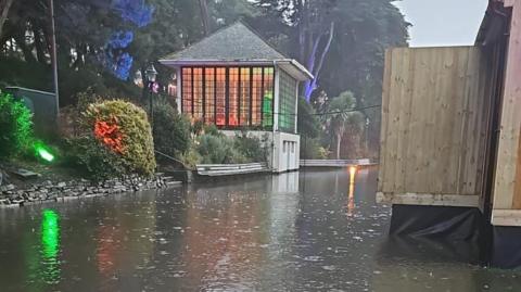 A flooded Bourne Stream with the bandstand and shrubs on the bank on the left. On the right is a raised wooden structure. The base is partly submerged.