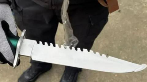 A large serrated knife is held by a police officer wearing grey-and-black protective gloves.