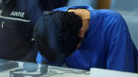 A Syrian national sits, his head bowed and wearing a blue T-shirt, behind a glss screen in a court room in Germany 