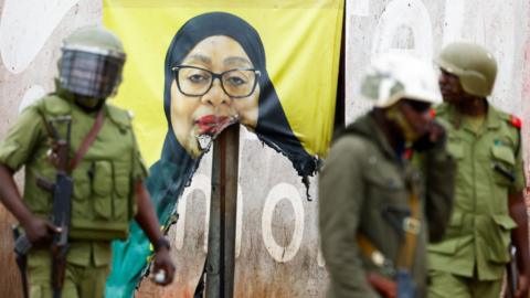 Tanzanian riot police officers (in jungle green uniform) walk past a vandalised campaign poster of President Samia Suluhu Hassan, following a protest a day after elections