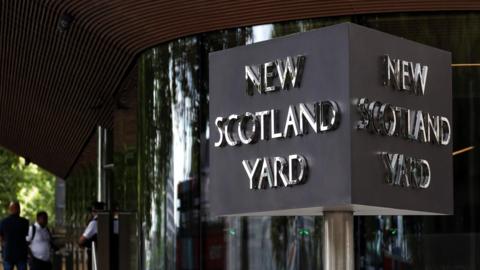 A sign outside Met police headquarters, with silver capital letters on a black background. It says 'New Scotland Yard'.