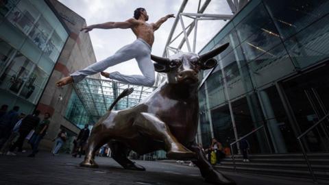 A male dancer wearing white trousers leaps high into the air next to a bull statue with glass buildings behind him