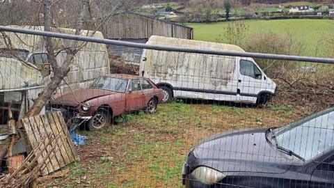 The view of the farm from a nearby road. Two white vans and a brown classic car can be seen in a bad condition behind a metal fence. Green fields and sheds can be seen behind them.