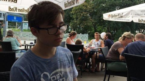 Thomas Parfett, a young man wearing a faded blue t-shirt and black-framed glasses. He is sat in what looks like an outside bar area, possibly abroad as there is German writing on signs behind him.