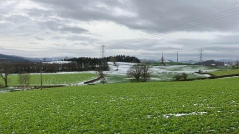 Green fields with a light dusting of snow on and in the background a few wind turbines and electric transmission towers can be seen. The sky is grey with many clouds.  