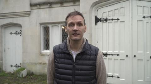 A man with greying hair and wearing a gillet over a long-sleeved shirt. He is standing outside a property.