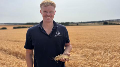 A man with ginger hair in a navy polo shirt. He's holding barley and stood on a field of barley. His shirt has 'manor farm, Sixpenny Handley ltd' embroidered on it.