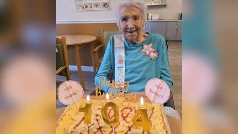 Meg, a woman in her hundreds, sat in front of a cake with candles reading the number "104". She is also wearing a birthday sash.