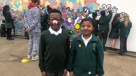 Two schoolchildren can be seen facing the camera, wearing comic relief noses that have had their own colours and designs drawn on. Schoolchildren can be seen in the background drawing on the mural with Mr Doodle, in his black and white suit, watching on.