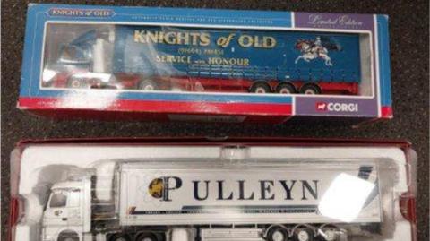 A shot from above of two toy lorries in their packaging. One is a blue Knights of Old lorry and the other is a white Pulleyn lorry.