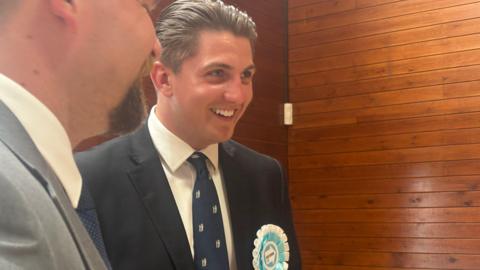 George Finch wears a Reform rosette and a navy tie with the Warwickshire bear and staff. He wars a dark suit jacket and a white shirt and has brown slicked back hair. He is pictures at the count for the local elections.