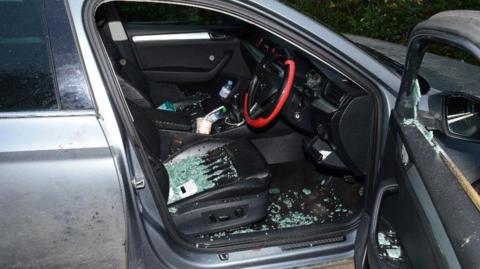 The driver's side of a car has a smashed window. The door is open and there is shattered glass all over the seat the the footwell. The steering wheel has a red rim
