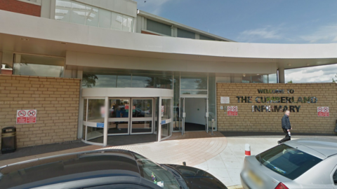 An exterior view of Cumberland Infirmary which is a pale brick building with two glass automatic doors, one which is slightly curved. The words 'Welcome to the Cumberland Infirmary' are written on the wall in silver letters. 