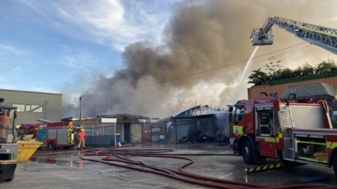 Smoke billows from a building with two fire engines and hose pipes in the foreground.