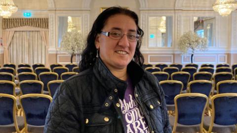 A woman with dark hair and glasses standing in a ballroom with glass chandeliers.