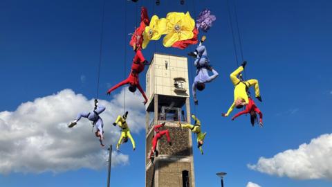 Aerialists wearing blue, yellow and red leotards suspended from ropes performing around some giant handmade flowers and a tower with a blue sky in the background 