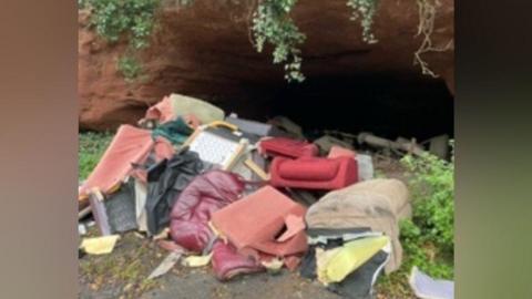 A pile of rubbish left on grass including parts of a red sofa, pink foam and black rubbish bags. There is the opening of a cave behind the fly tipping 