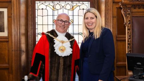 Steven Crellin, wearing a red robe with a mayoral chain, stands alongside former mayor, Natalie Byron-Teare, a woman with blonde hair dressed in a navy blue suit. Wooden panelled walls and a decorative stained-glass window can be seen in the background.