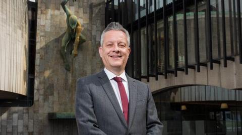 Nick Kemp standing in front of the Newcastle Civic Centre. He has grey spiky hair and is wearing a grey suit and red tie.