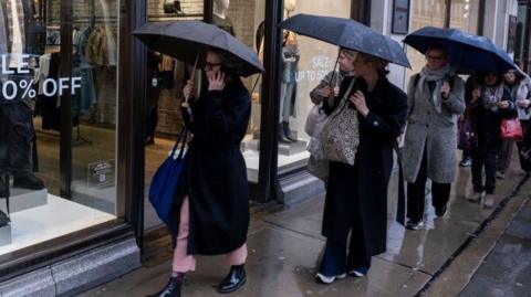 Women walking past a shop window carrying black umbrellas on a rainy day