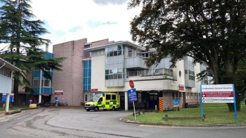 The exterior of Cheltenham General Hospital, where an ambulance is parked outside. There are three floors, with the bottom being a garage-like structure. There are blue design elements on the building which looks quite dated. There are some large coniferous trees and a lawn space, along with signage that reads "Cheltenham General Hospital" along with sign posts for various departments within the hospital.