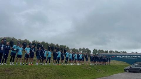 Staff and players line up on a grass verge outside Carlisle United's Brunton Park stadium