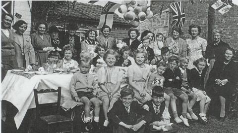 A black and white photo showing a group of people, mainly children, in 1950s clothing, sat around a table smiling at the camera, with union flags hanging in the background.