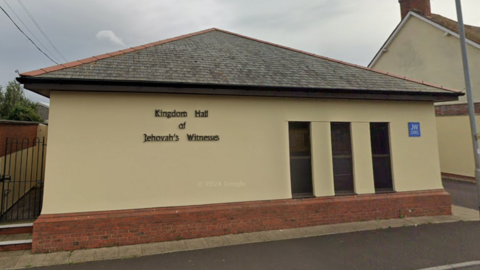 The Kingdom Hall, which is a one-storey building rendered with a pale yellow plaster. It has three small windows, a sign which says "Kingdom Hall of Jehovah's Witnesses" and a blue plaque which says JW.org. 