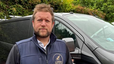 There is a man with brown brushed-up hair and a brown beard. He is wearing a long-sleeve black shirt with a dark blue gilet with 'A.J Harrison Painter & Decorator in yellow text in a badge. He is stood infront of his black work van