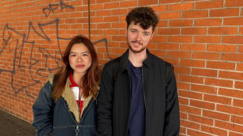 Two young people stand close together beside a long brick wall marked with graffiti. They are wearing trendy modern clothing. The woman on the left has red hair. The man on the right has brown hair and a beard. 