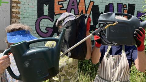 Three women standing in front of a brick wall. The woman in the middle is holding a black bag over her face and those to either side of her are holding plastic watering cans over their faces.