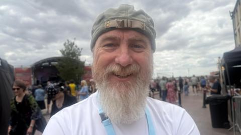 Mark Page - a man in a grey cap with a long grey beard and moustache is smiling into the camera lens. He is wearing a white top, blue lanyard and you can see a stage and people dotted about on a street behind him.