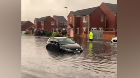 A car is submerged up to it's number plate in flood water on a residential road. Red houses can be seen bordering the houses.