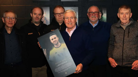 A group of six men are standing in a line. The man in the middle John Richards is holding a board bearing Diogo Jota's image with writing at the bottom
