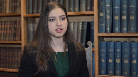 Arwa Elrayess is standing in front of old bookshelves. She has long dark hair and is wearing a dark blazer over a green jumper.