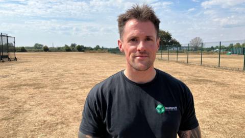 Andrew Briggs stands on a dried out sports pitch. The grass is brown and yellow after weeks of drought. He is wearing a black T shirt with a small green symbol on it, and has short dark brown hair