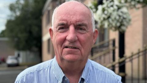 A older man looks into the camera. His hair is white around the side of his head and he is bald on top. He is wearing a blue and white striped shirt.