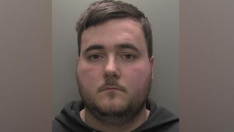 A police custody shot of a man with short brown hair and brown eyes. He has a thin stubbly beard and is wearing a black hooded sweatshirt.