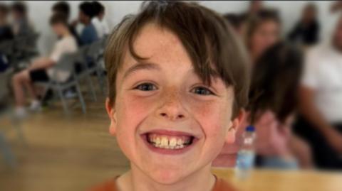 A boy called Rex is smiling at the camera - there are some people in the background of the photo