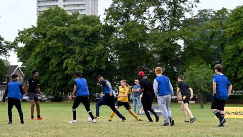 Children, some wearing blue bibs, play football in a park