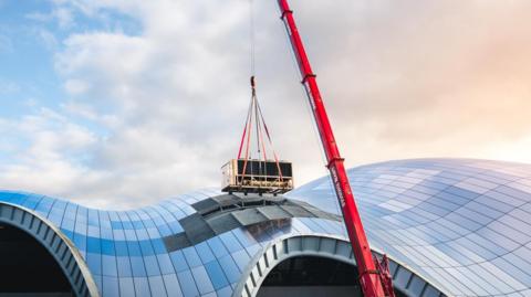 A large chiller unit is strapped to a red crane and lifted out of The Glasshouse where glass panels have been removed from the roof of the building. The orange, blue and grey sky is reflected on the curved glass roof of The Glasshouse.  