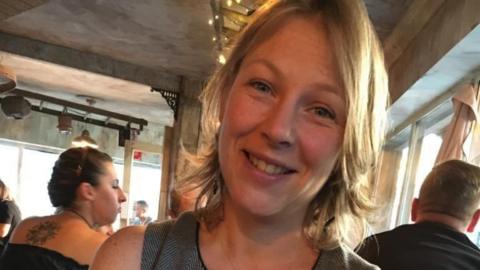 A head and shoulders picture of Alison Clark, smiling for the camera at a social event. She has shoulder-length, blonde hair and is wearing a grey vest-top.