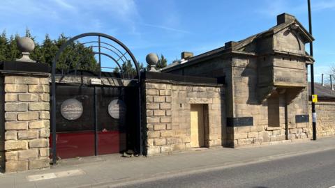 The guardhouse on Barrack Road. The bulding is made of large sandstone bricks. It's doors and windows have been boarded up. The entranceis made of red tinted glass and displays signs for a now closed Lebanese restaurant.